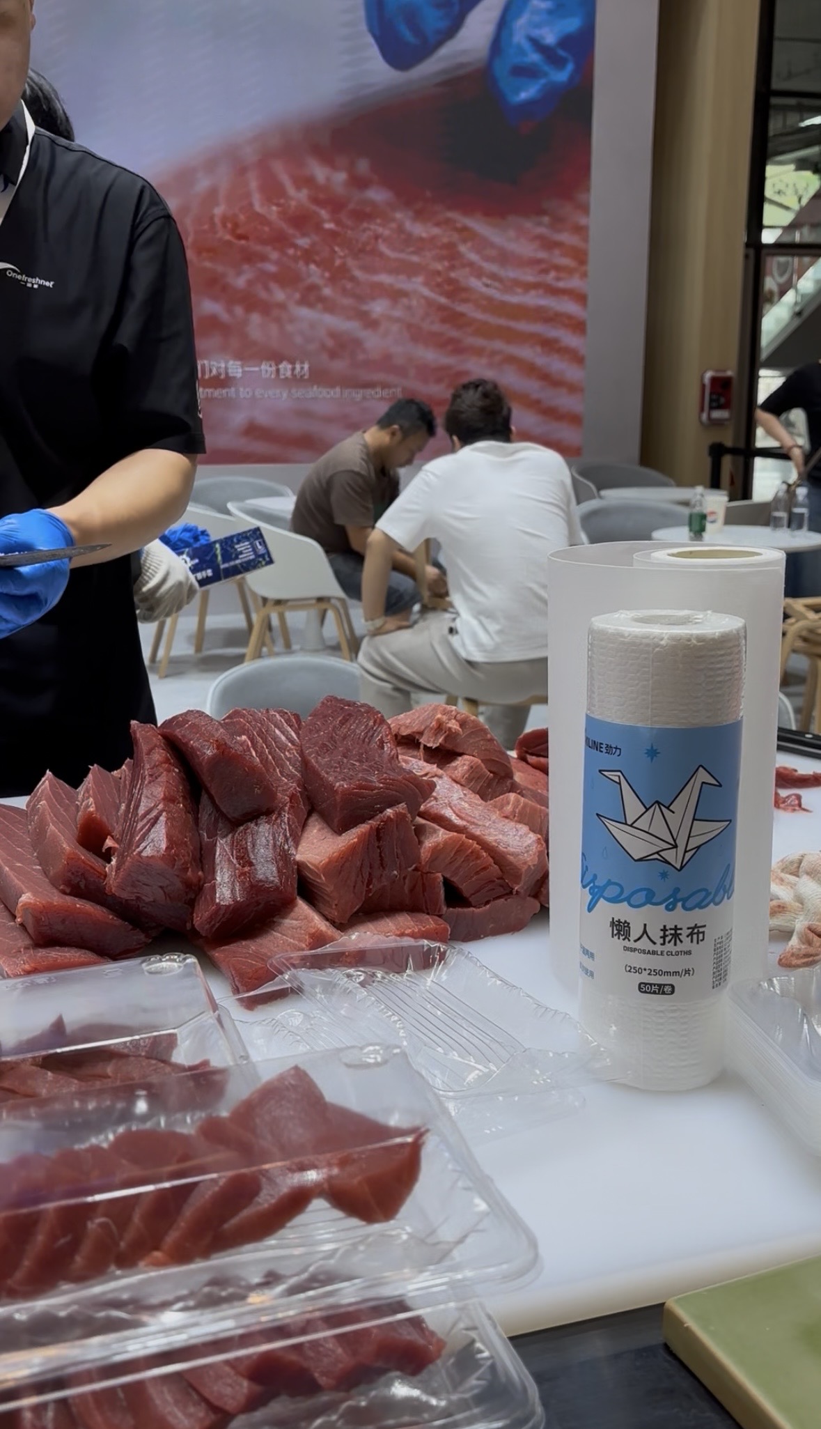 渔博会现场食品企业使用KILINE懒人抹布处理新鲜金枪鱼:操作员身穿黑色工作服、戴蓝色丁腈手套,案台摆放保鲜金枪鱼块与卷装懒人抹布
